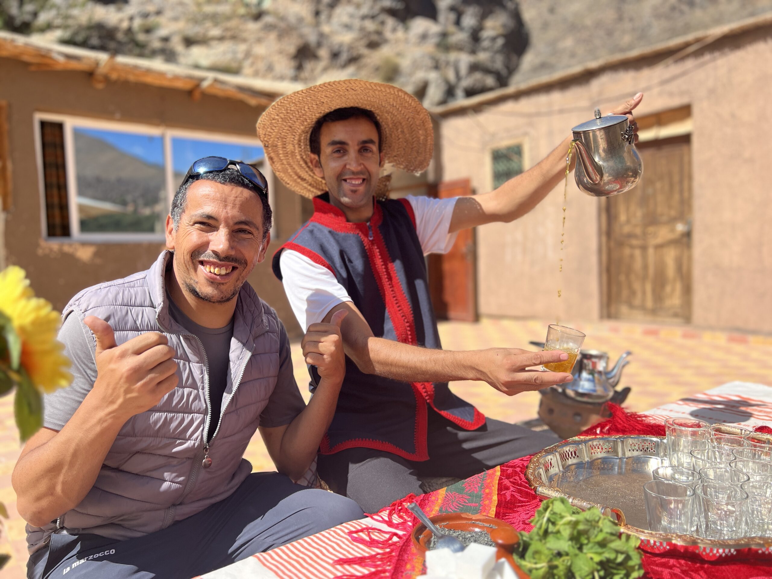 A man with thumbs-up sits next to another who is pouring tea in a arid Moroccan village.