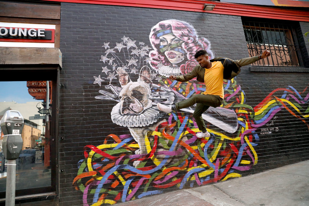 Serge Jr, a San Francisco based Artist, is jumping in the air, in a yellow, shirt, in front of one of his colorful murals of a dog in a ballerina outfit.