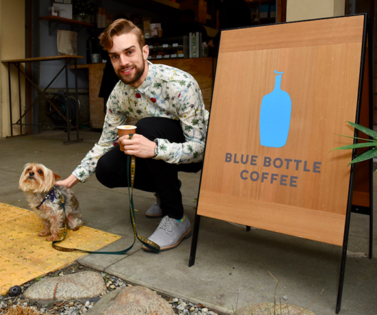 Myles Thatcher poses with a yorkie in front of a sign for Blue Bottle Coffee.
