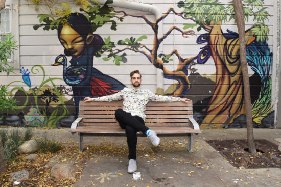Myles Thatcher poses in a colorful top while sitting on a bench across from Blue Bottle coffee in Hayes Valley, San Francisco. There is a beautiful mural in the background.