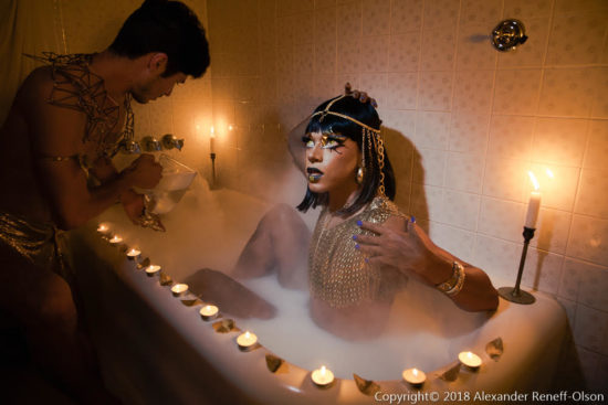 A drag queen poses in Egyptian attire, in a bathtub, with an assistant photographing her.
