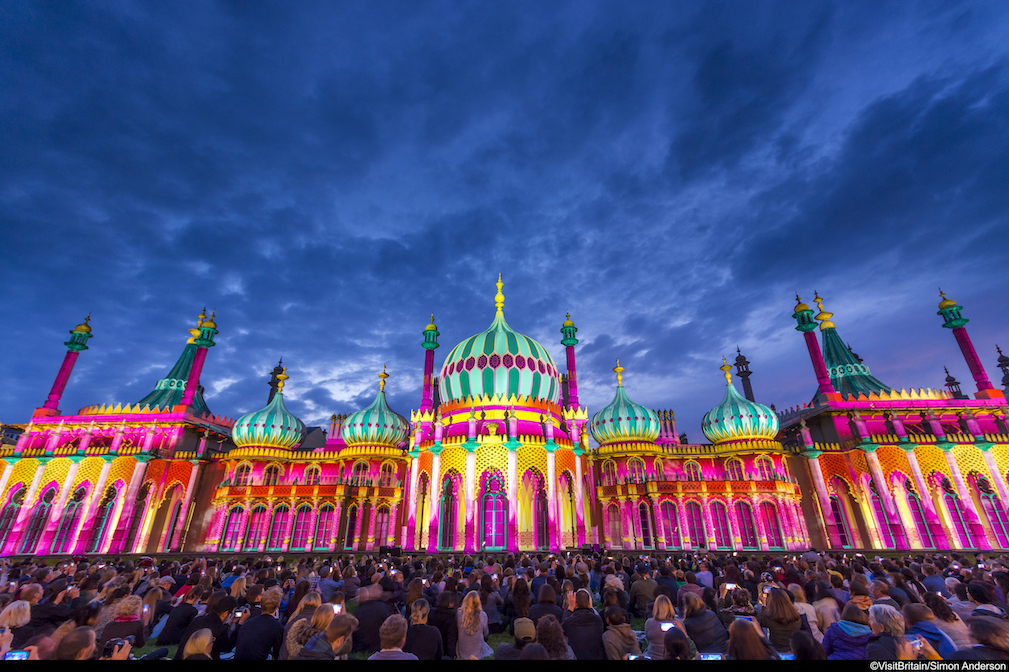 Crowd of people in front of the Royal Pavilion at dusk, lit up in a range of vivid colours, pink, green, yellow and purple, Brighton, East Sussex, England.