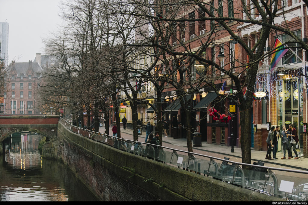 Distant view of group of gay men having drinks outside in Manchester. River and and tree lined street in view. Gay flag.